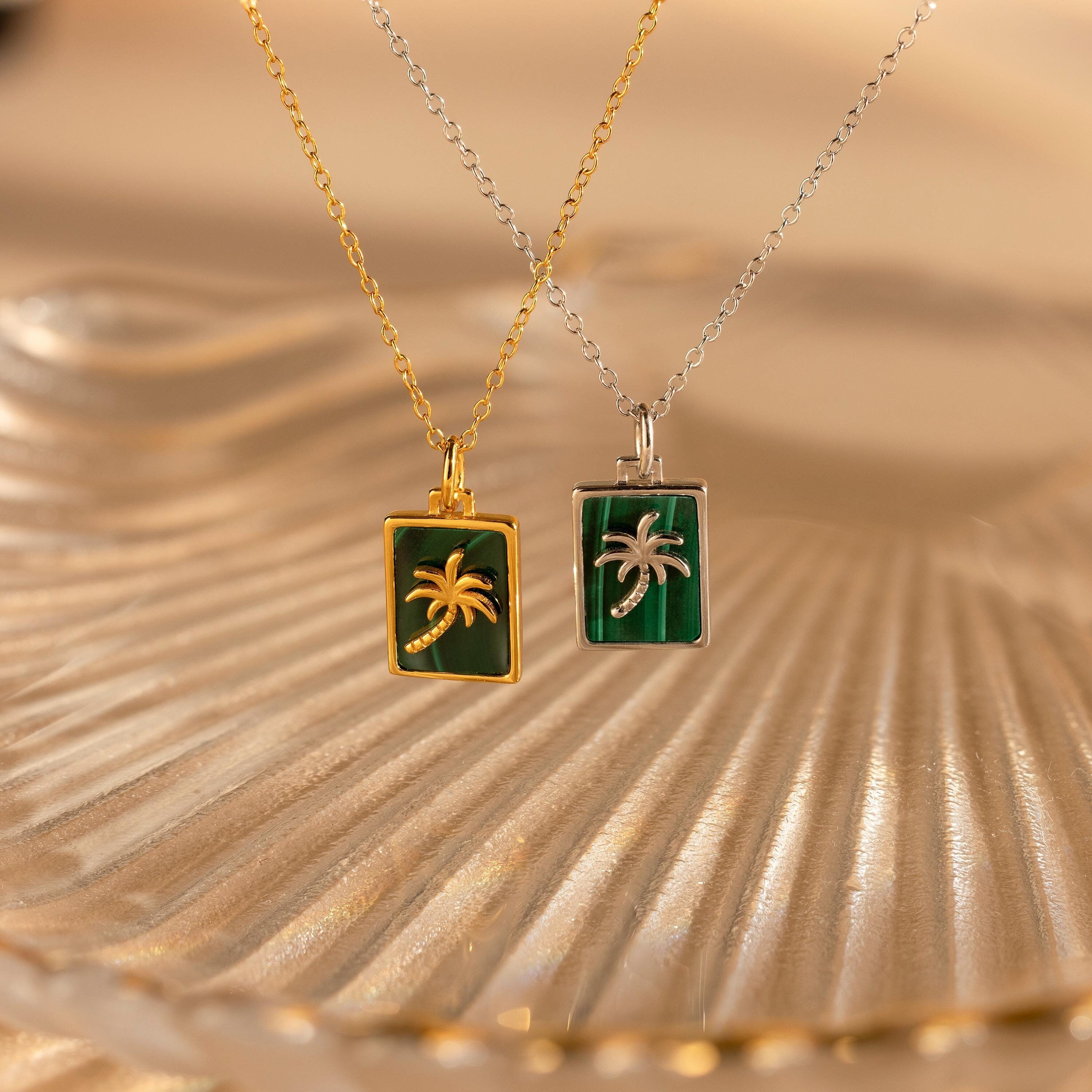 Two Malachite Palm Tree Necklaces—one gold and one silver— from our collection, displayed on textured glass.