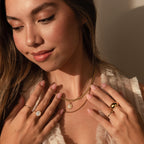 Woman with long brown hair wears layered gold necklaces, including the delicate Pearl Tulip Necklace, along with rings, as she looks down and smiles softly.