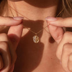 A person holds the Pearl Tulip Necklace, featuring a gold oval pendant with a delicate flower design, close to their neck.