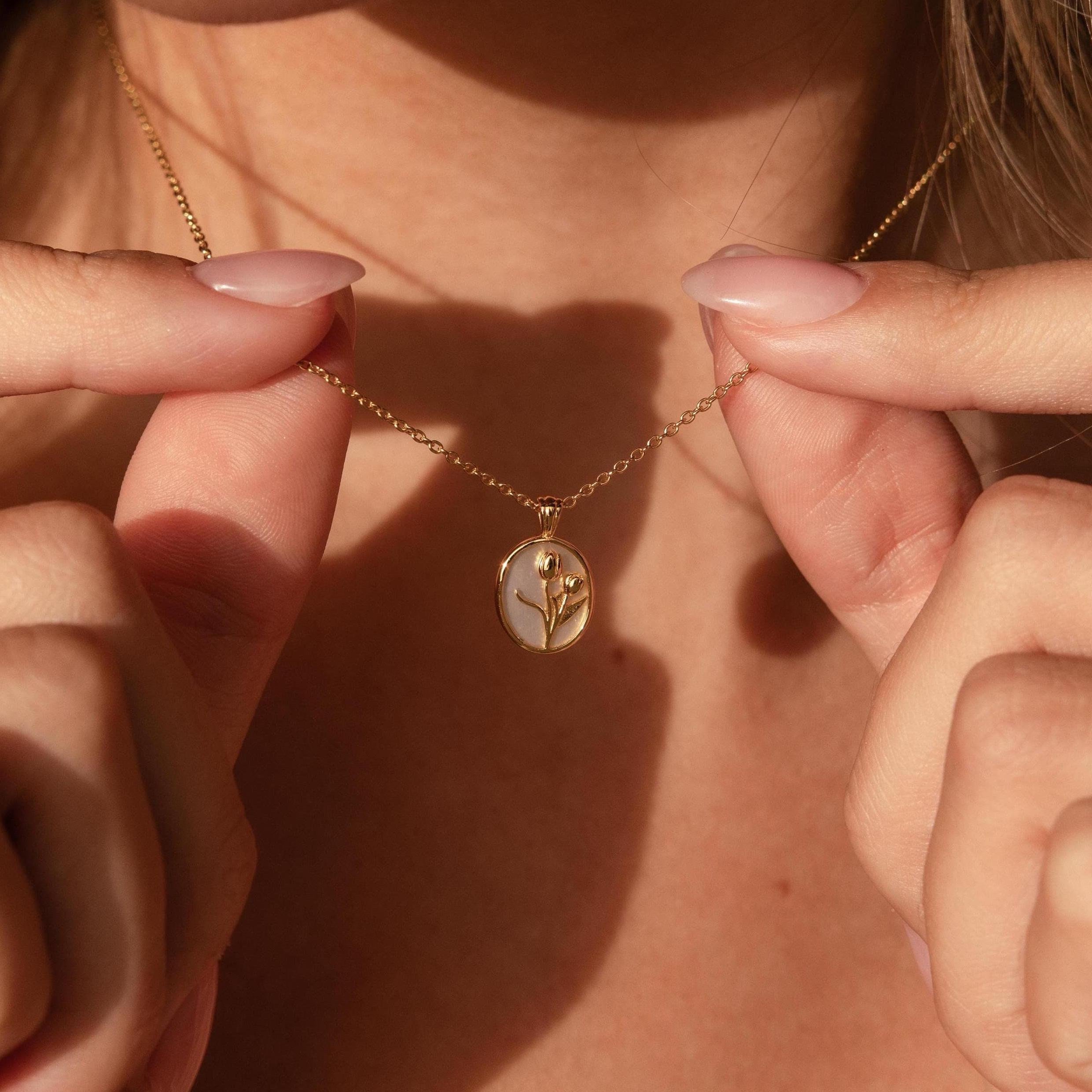 A person holds the Pearl Tulip Necklace, featuring a gold oval pendant with a delicate flower design, close to their neck.