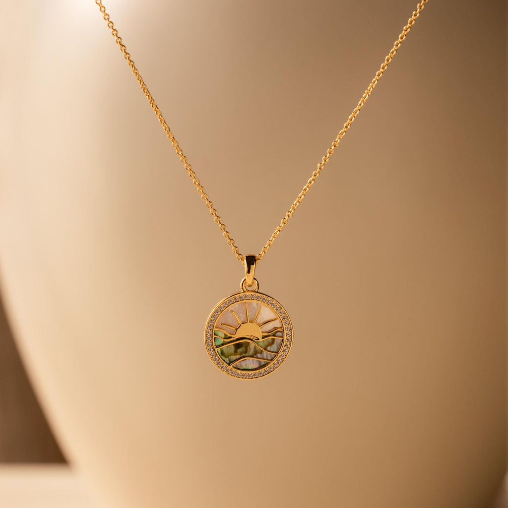 The Abalone Sunrise Pendant Necklace in 18K Gold features a round sun and landscape design, reminiscent of an abalone sunrise, displayed against a soft beige background.