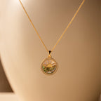 The Abalone Sunrise Pendant Necklace in 18K Gold features a round sun and landscape design, reminiscent of an abalone sunrise, displayed against a soft beige background.