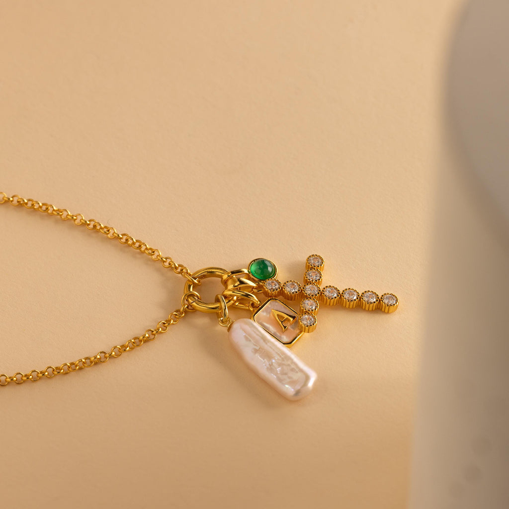 Gold Pave Cross Charm is paired with a gold rolo necklace, long pearl charm, emerald birthstone charm and initial charm