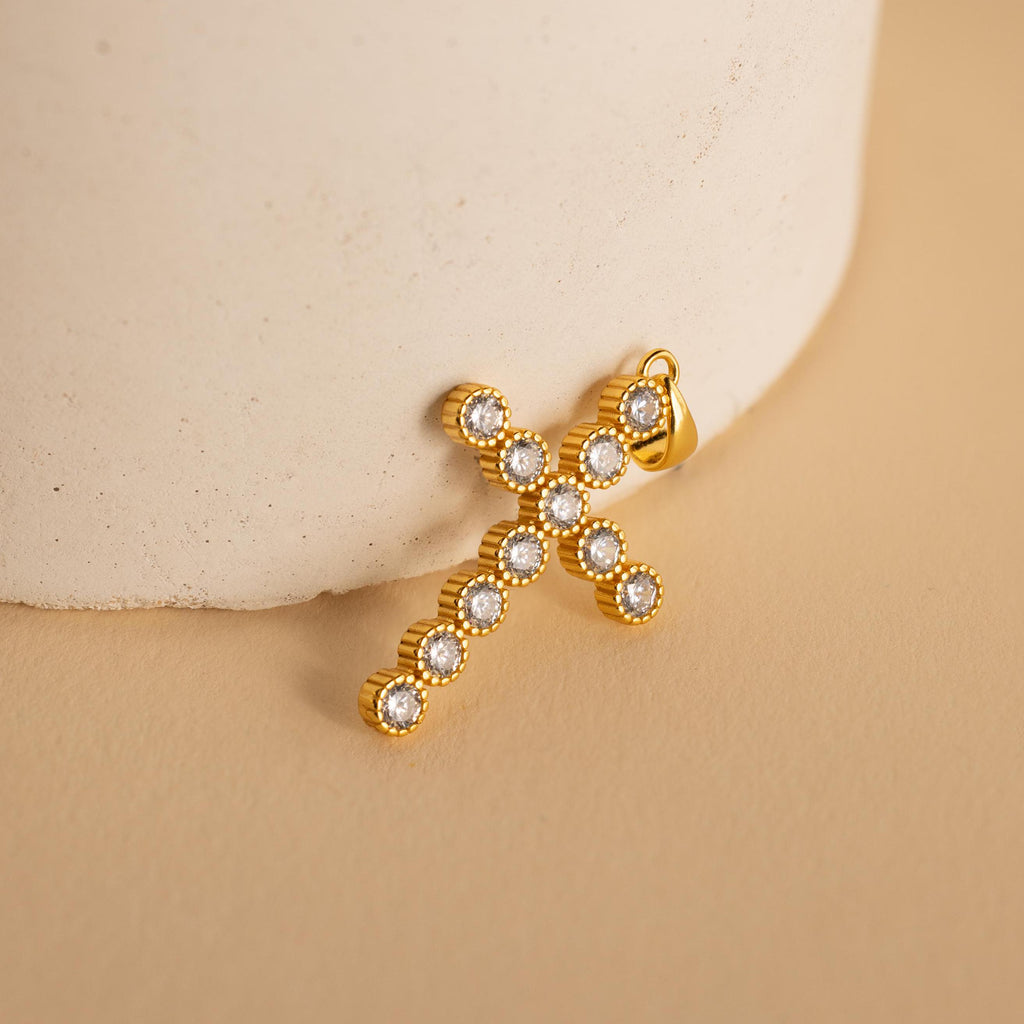 Pave Cross Charm in 18K Gold leaning on a vase. The charm is created with multiple round 4mm CZ Diamonds arranged in the shape of a cross with a ribbed bezel setting. 