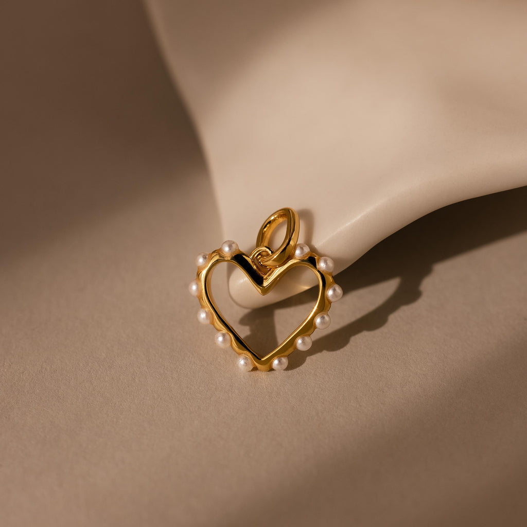 The Classic Pearl Heart Charm in 18K Gold, a heart-shaped pendant adorned with small pearls, rests on a smooth, light-colored surface.