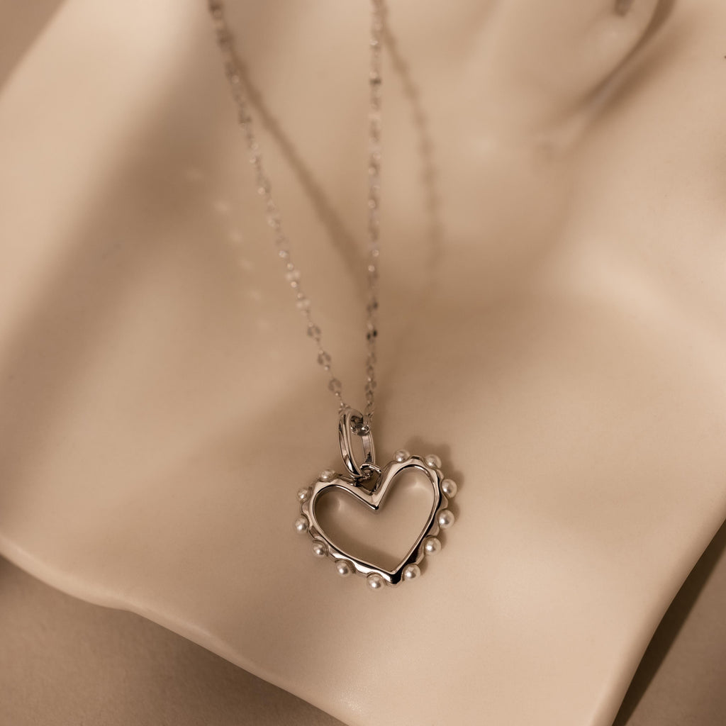 The Classic Pearl Heart Charm in Sterling Silver features pearl beads and a delicate heart pendant, beautifully displayed on a soft beige surface.