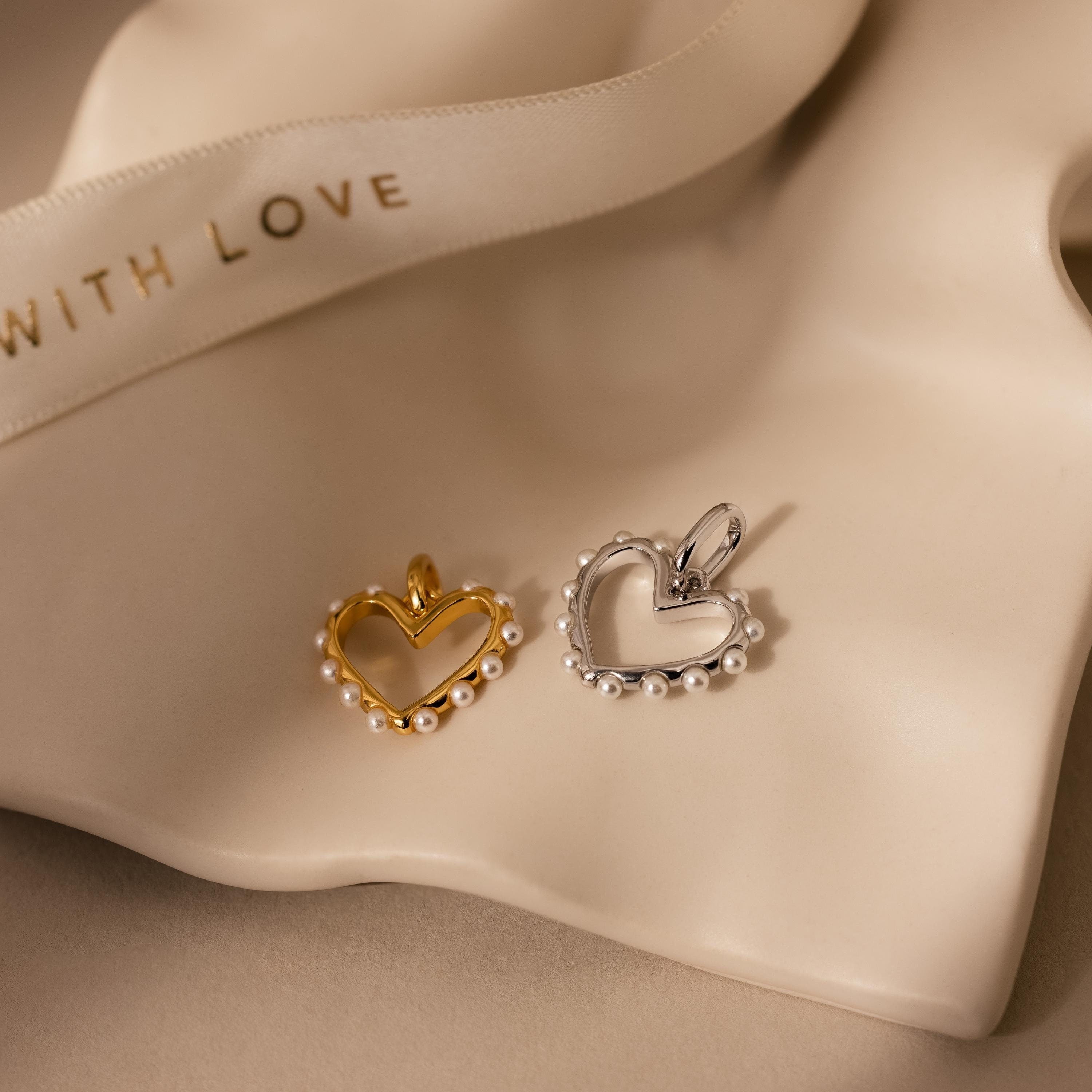 The Classic Pearl Heart Charm, a gold and silver heart pendant with pearls, rests on cream fabric beside a ribbon that reads WITH LOVE.
