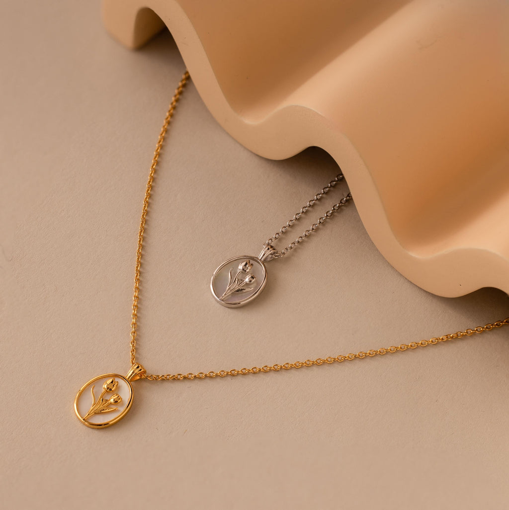 The Pearl Tulip Necklace in Sterling Silver features a delicate floral pendant, showcased on a beige surface beside a wavy ceramic object.