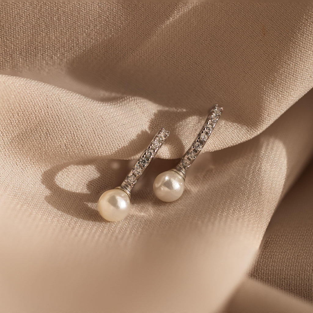 The Pave Pearl Drop Huggies, featuring sparkling diamonds and luminous pearls, are showcased elegantly on beige fabric.