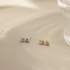 Two pairs of Pearl Aquamarine Studs, one in silver and one in gold, with blue aquamarine gemstones, rest on a beige surface by a glass—elegant and timeless earrings to elevate any look.