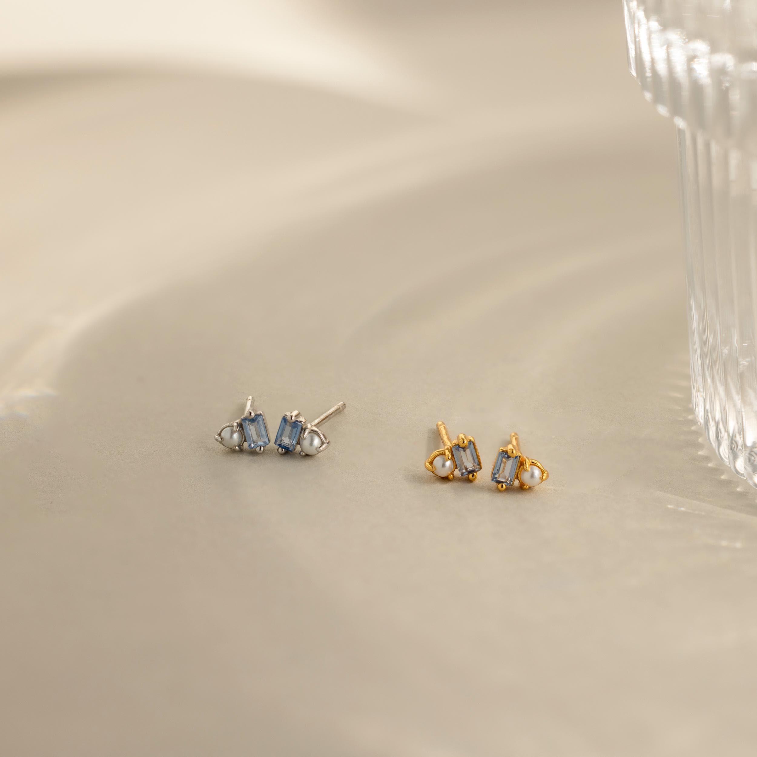 Two pairs of Pearl Aquamarine Studs, one in silver and one in gold, with blue aquamarine gemstones, rest on a beige surface by a glass—elegant and timeless earrings to elevate any look.