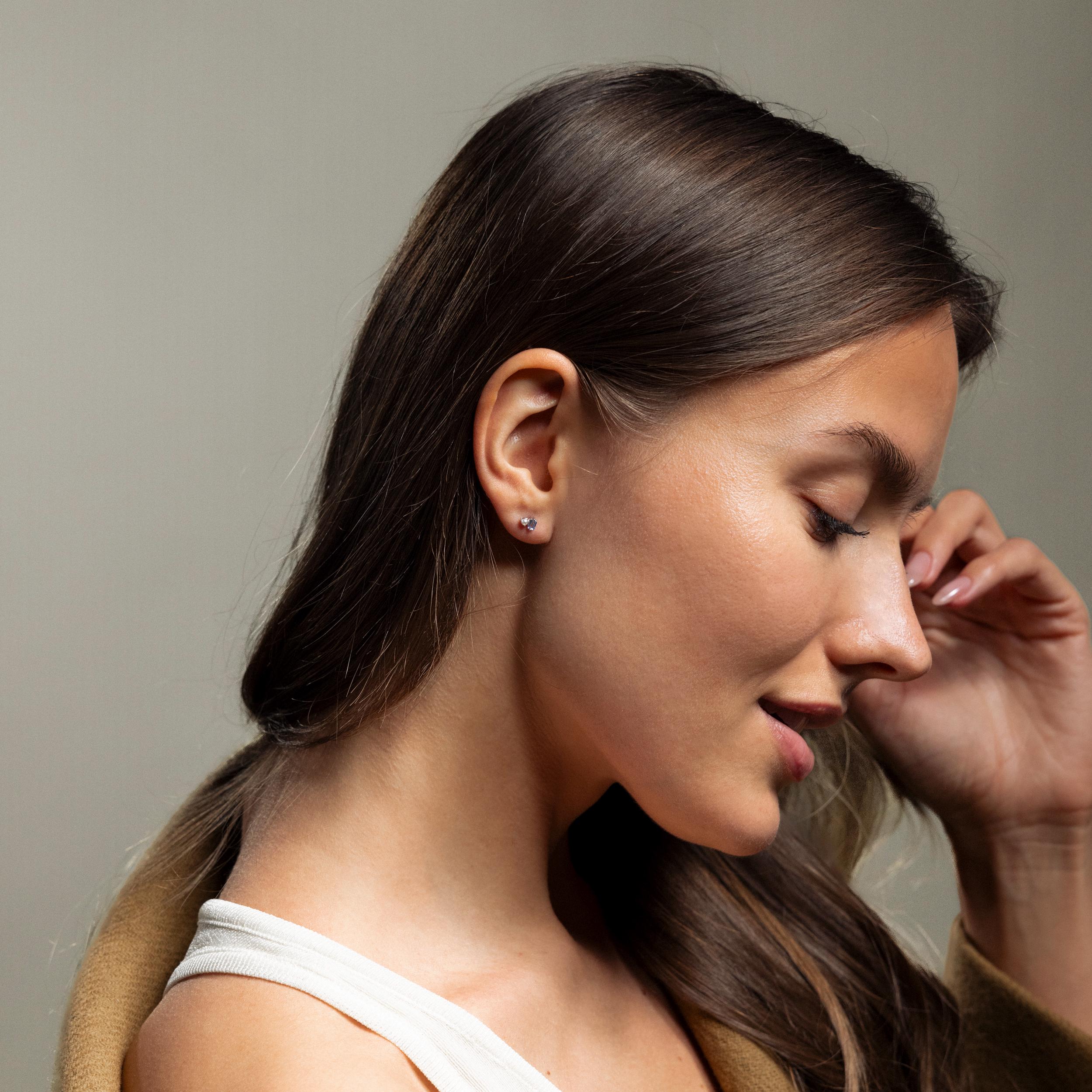 A woman with long brown hair, wearing a white top and Pearl Aquamarine Studs, looks down in soft lighting.