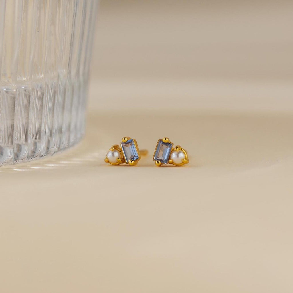 Pearl Aquamarine Studs, gold earrings featuring clear baguette gems and small pearls, are displayed near a ribbed glass on a beige surface.