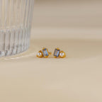 Pearl Aquamarine Studs, gold earrings featuring clear baguette gems and small pearls, are displayed near a ribbed glass on a beige surface.