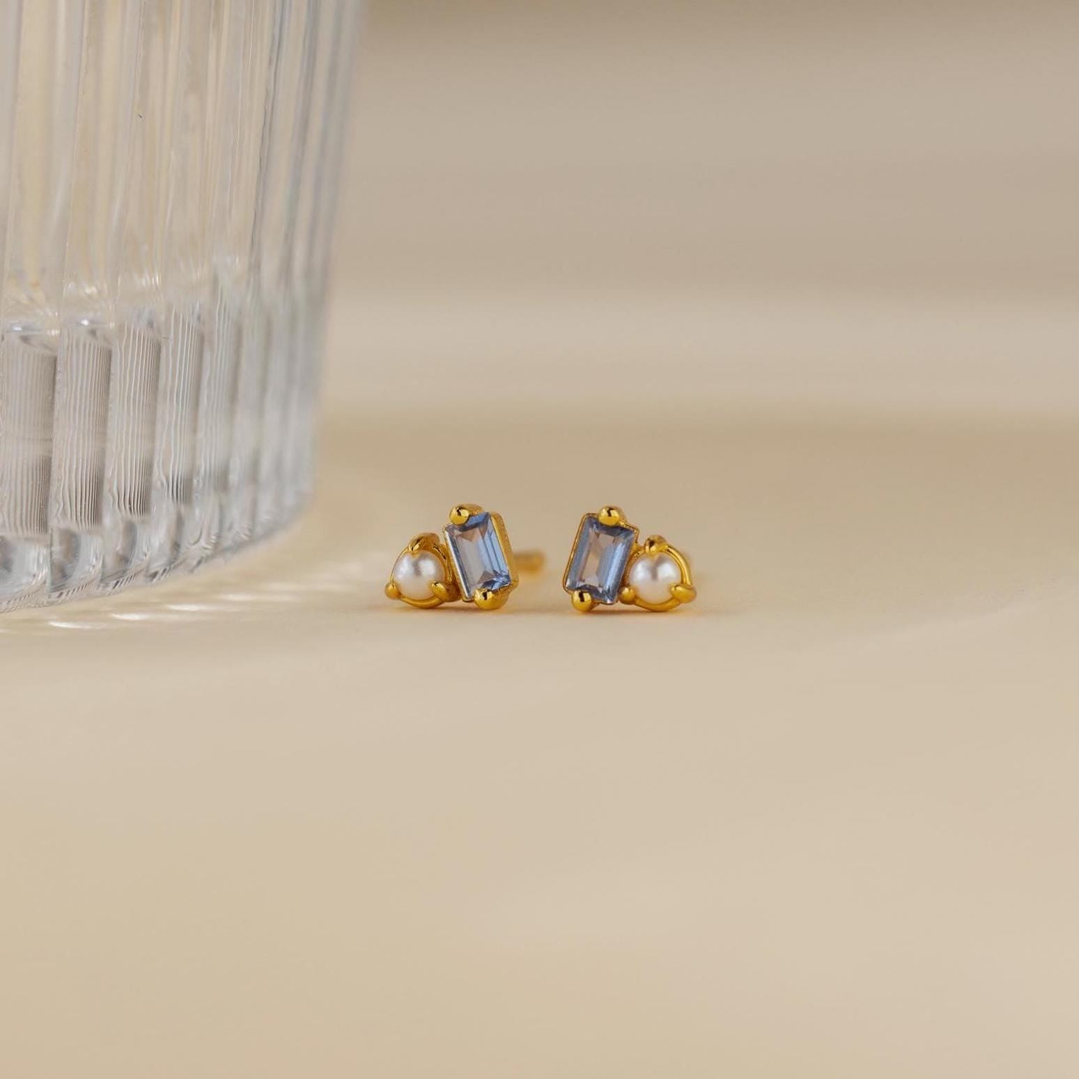 Pearl Aquamarine Studs, gold earrings featuring clear baguette gems and small pearls, are displayed near a ribbed glass on a beige surface.