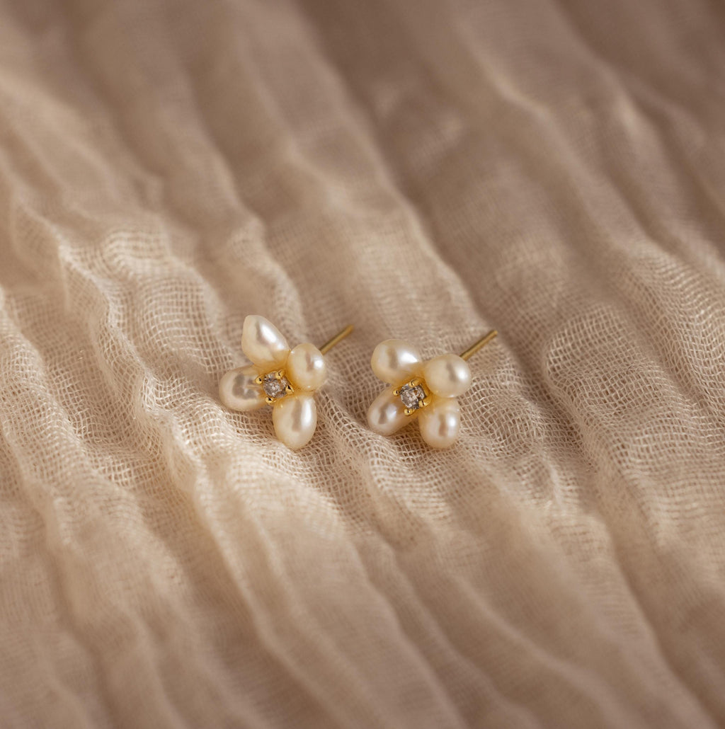 Pearl Flower Studs feature clusters of pearls and small gemstones in a floral design, beautifully displayed on textured beige fabric.