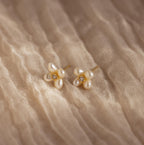 Pearl Flower Studs feature clusters of pearls and small gemstones in a floral design, beautifully displayed on textured beige fabric.
