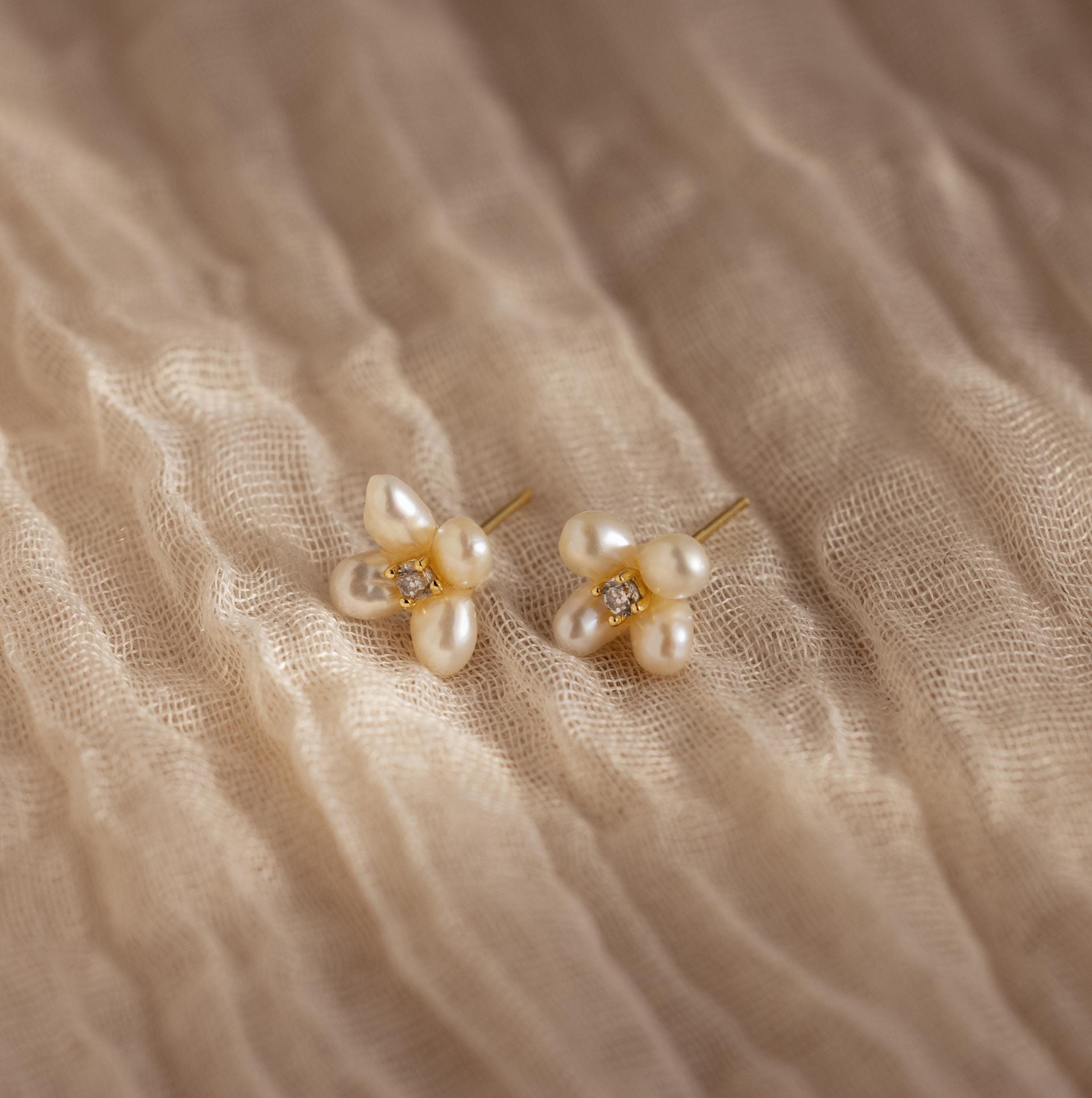 Pearl Flower Studs feature clusters of pearls and small gemstones in a floral design, beautifully displayed on textured beige fabric.