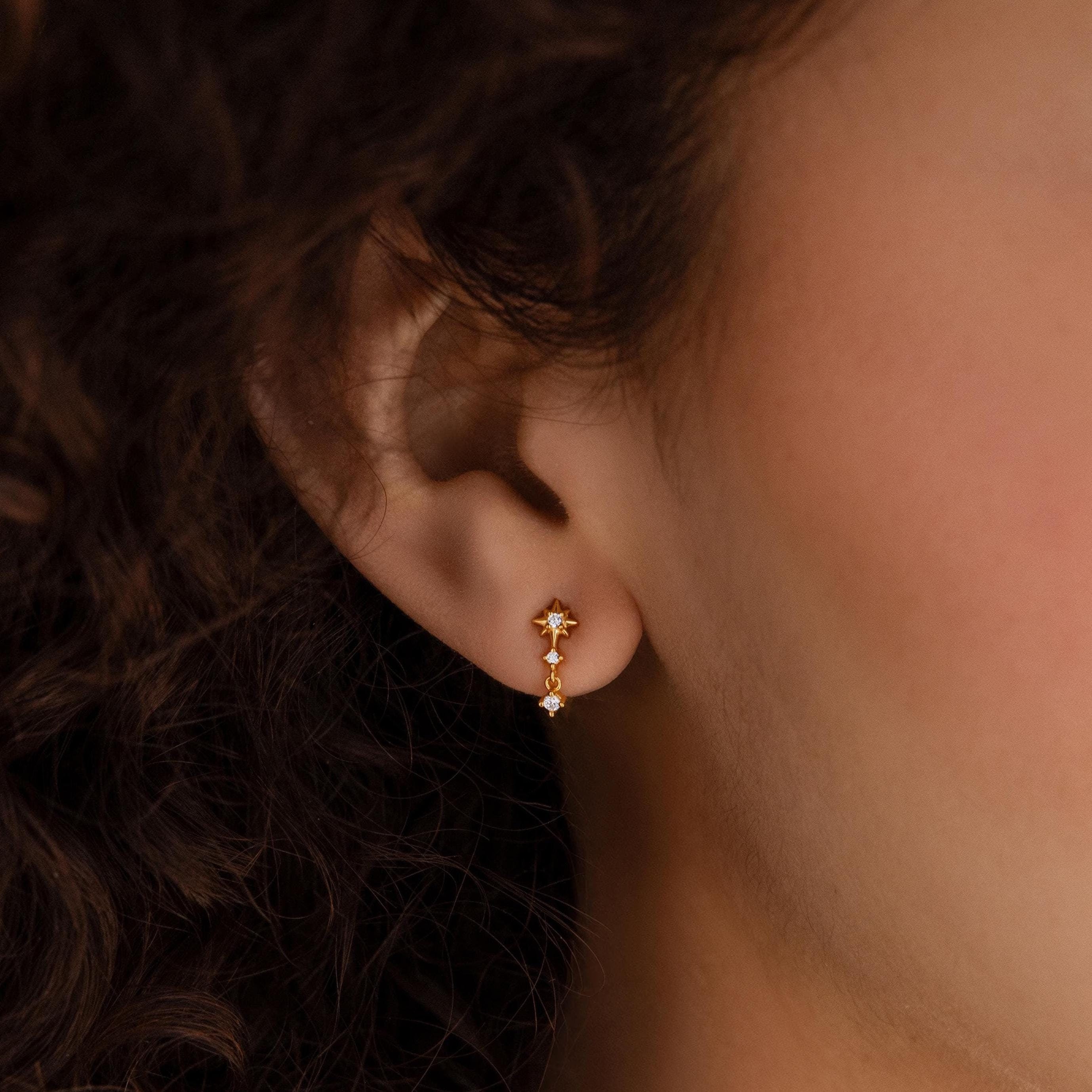 Close-up of a person’s ear wearing Diamond Starburst Drop Studs in 18K Gold, featuring two dangling star-shaped charms that shimmer like diamonds.