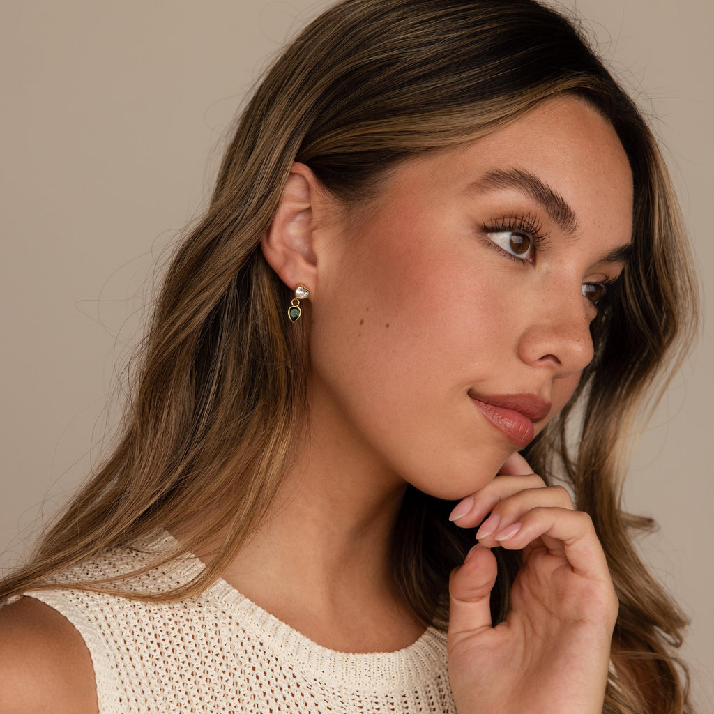 Woman with long hair wearing a cream knit top, looking to the side and touching her chin, styled with elegant Emerald Heart Drop Studs.