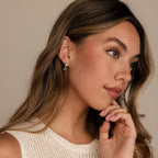 Woman with long hair wearing a cream knit top, looking to the side and touching her chin, styled with elegant Emerald Heart Drop Studs.