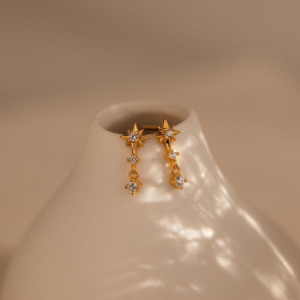 Diamond Starburst Drop Studs—gold star-shaped earrings with dangling crystals—are displayed on a smooth, off-white ceramic vase.