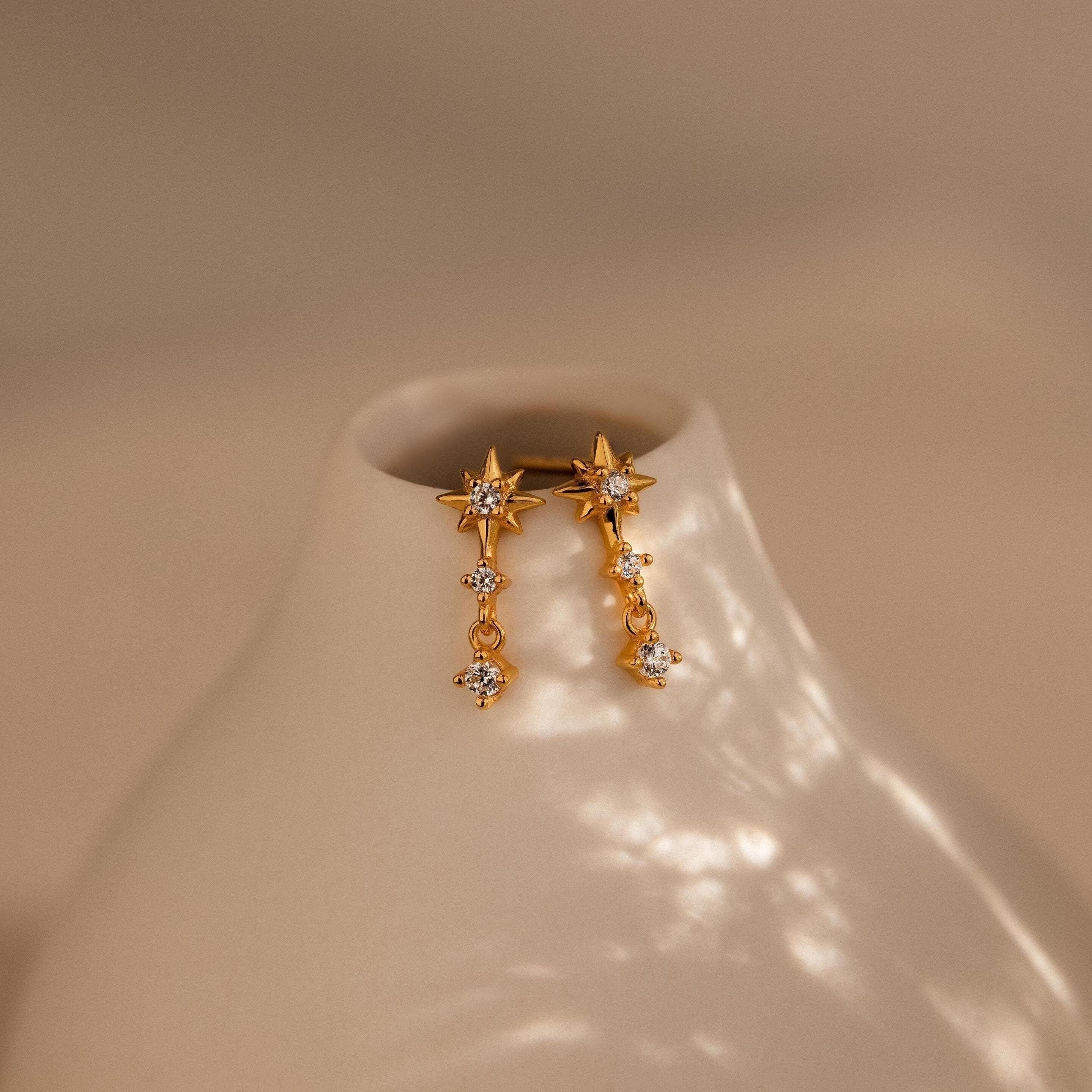 Diamond Starburst Drop Studs—gold star-shaped earrings with dangling crystals—are displayed on a smooth, off-white ceramic vase.