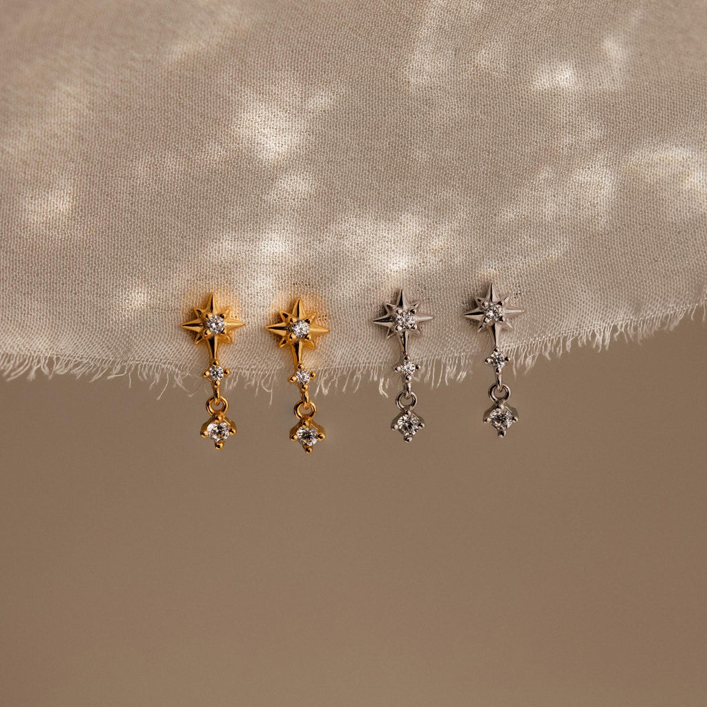 Two pairs of Diamond Starburst Drop Studs, one gold and one silver, are elegantly displayed on textured beige fabric.