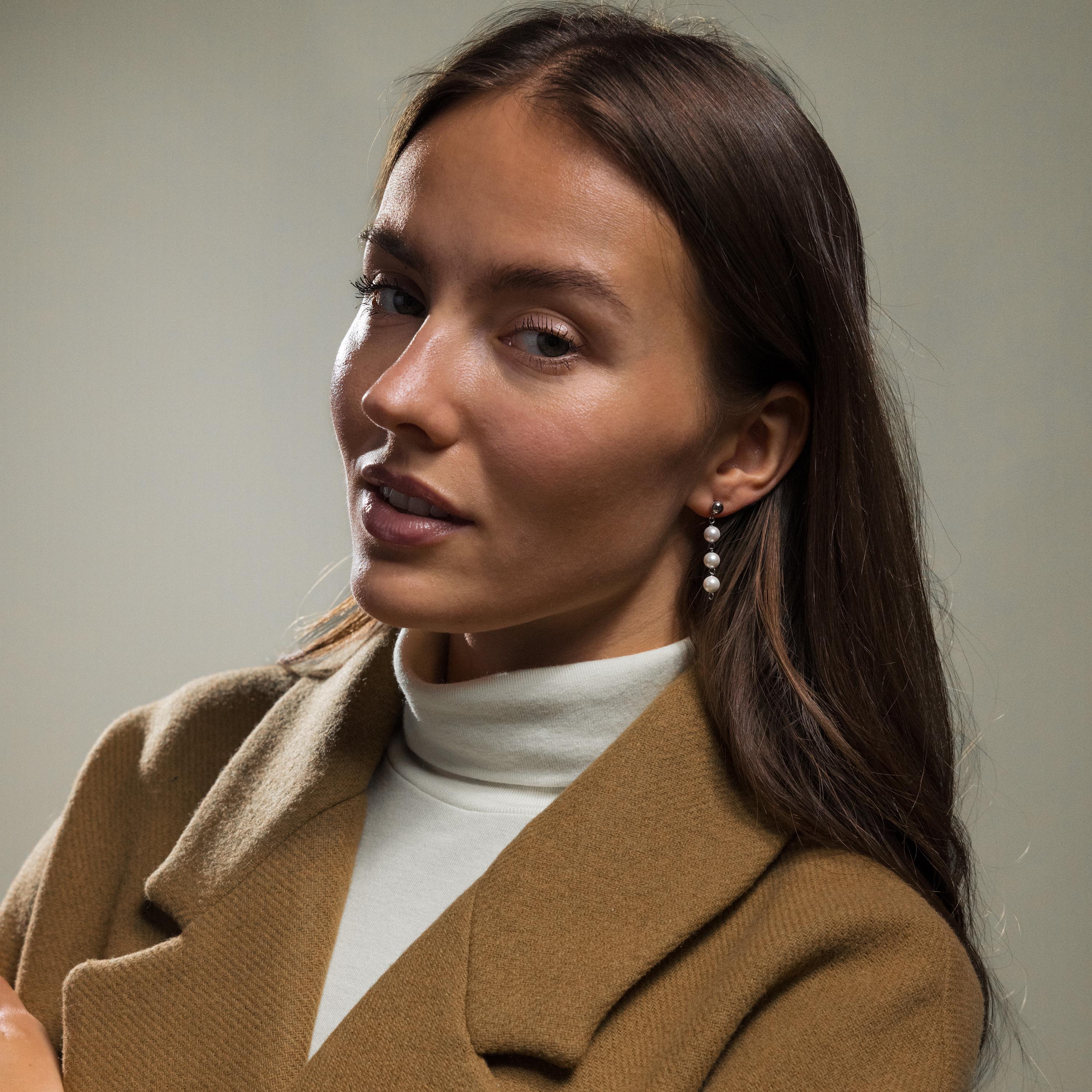 A woman with long brown hair poses calmly, wearing a brown coat, white turtleneck, and Dainty Pearl Drop Studs.