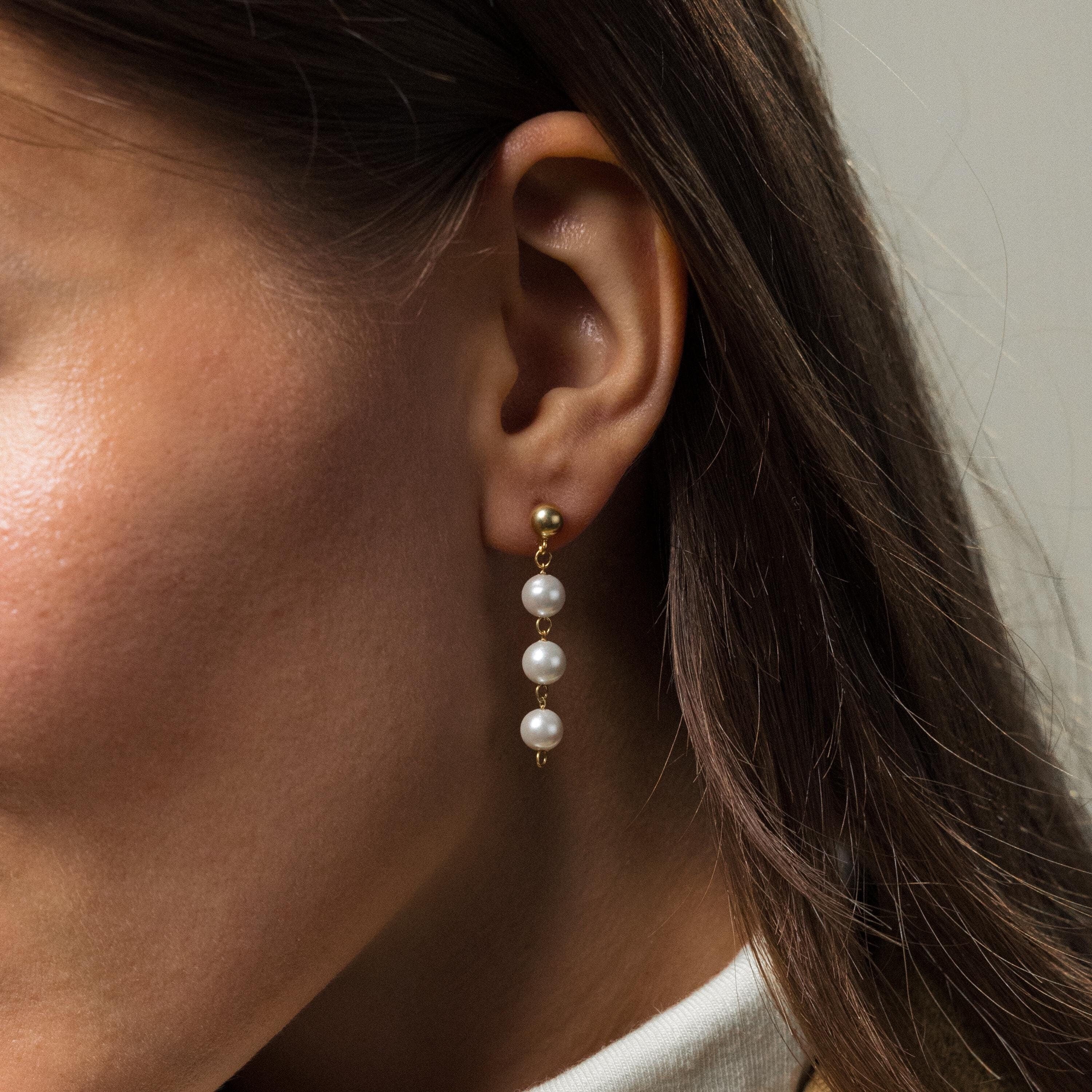 A close-up of a woman's ear wearing the Dainty Pearl Drop Studs, featuring three dangling white pearls in a delicate gold setting.