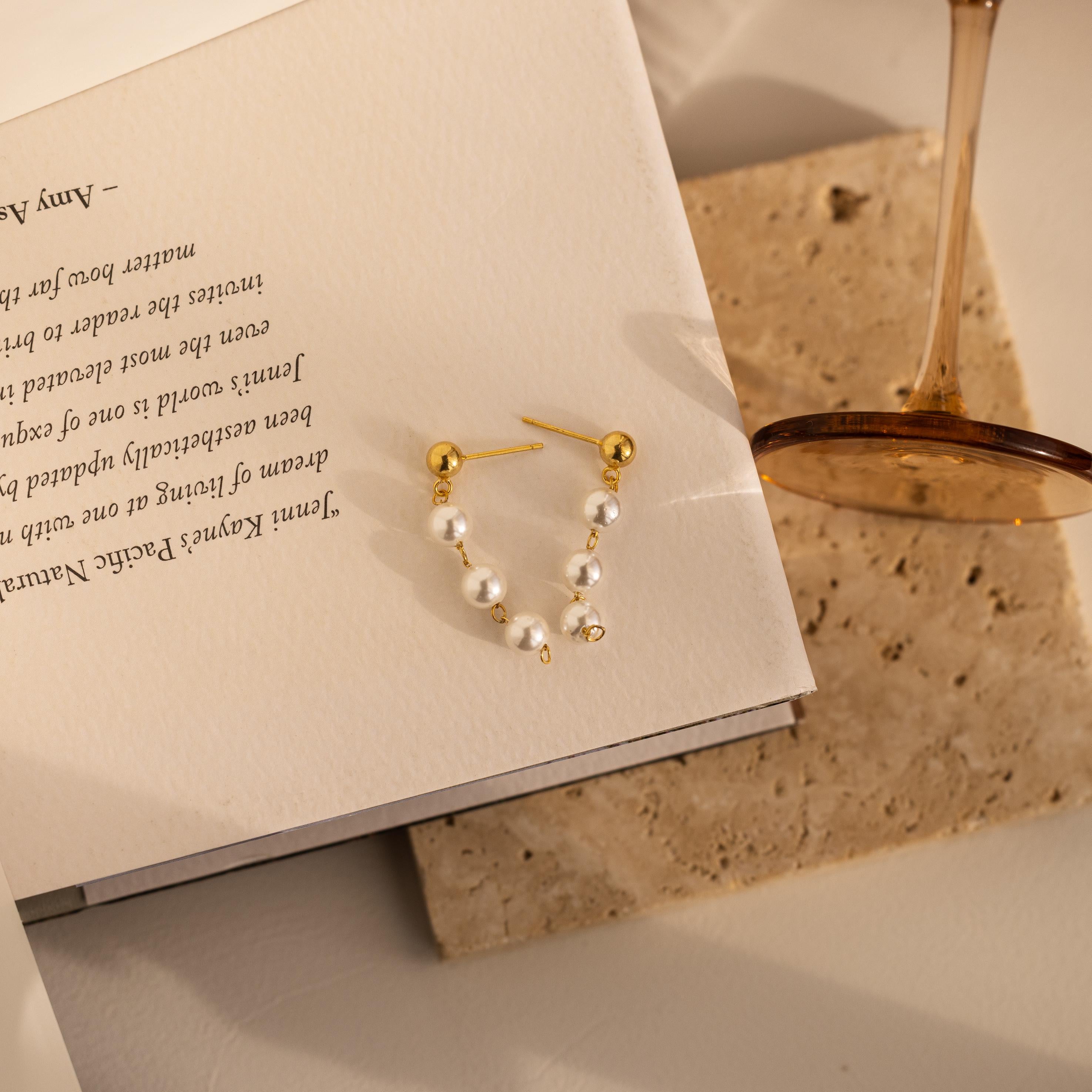 Dainty Pearl Drop Studs are shown resting on an open book beside a brown-stemmed glass atop a stone surface.