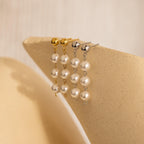Two pairs of Dainty Pearl Drop Studs—one with gold, one with silver—are displayed on a beige textured surface, highlighting their elegant pearl design.
