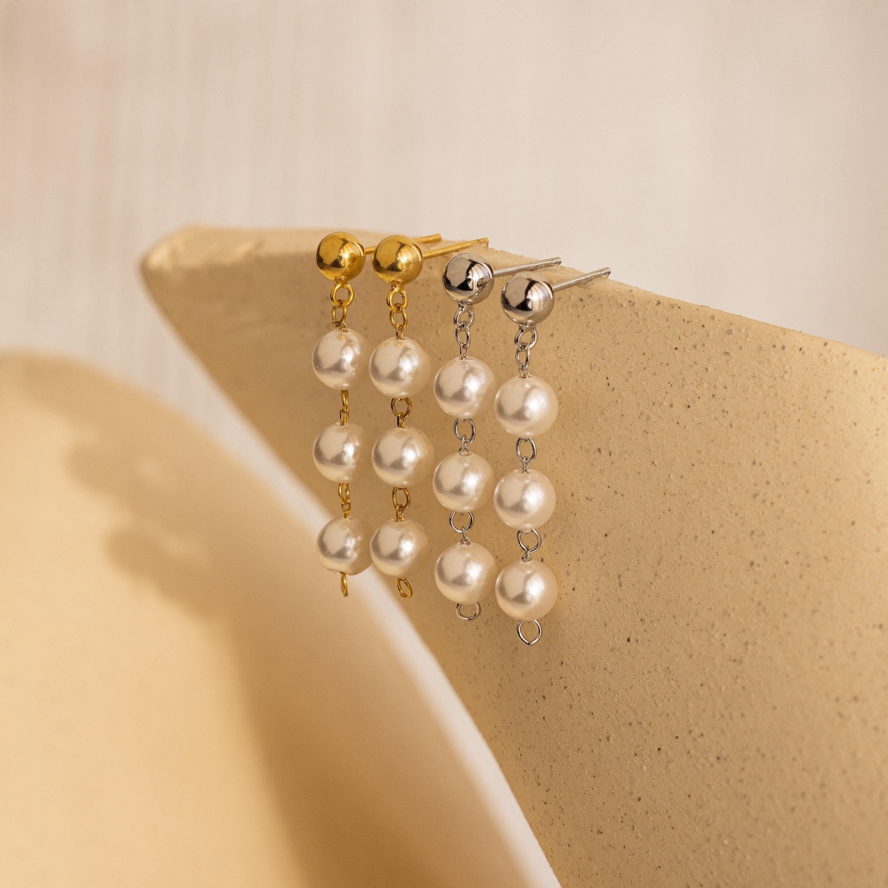 Two pairs of Dainty Pearl Drop Studs—one with gold, one with silver—are displayed on a beige textured surface, highlighting their elegant pearl design.