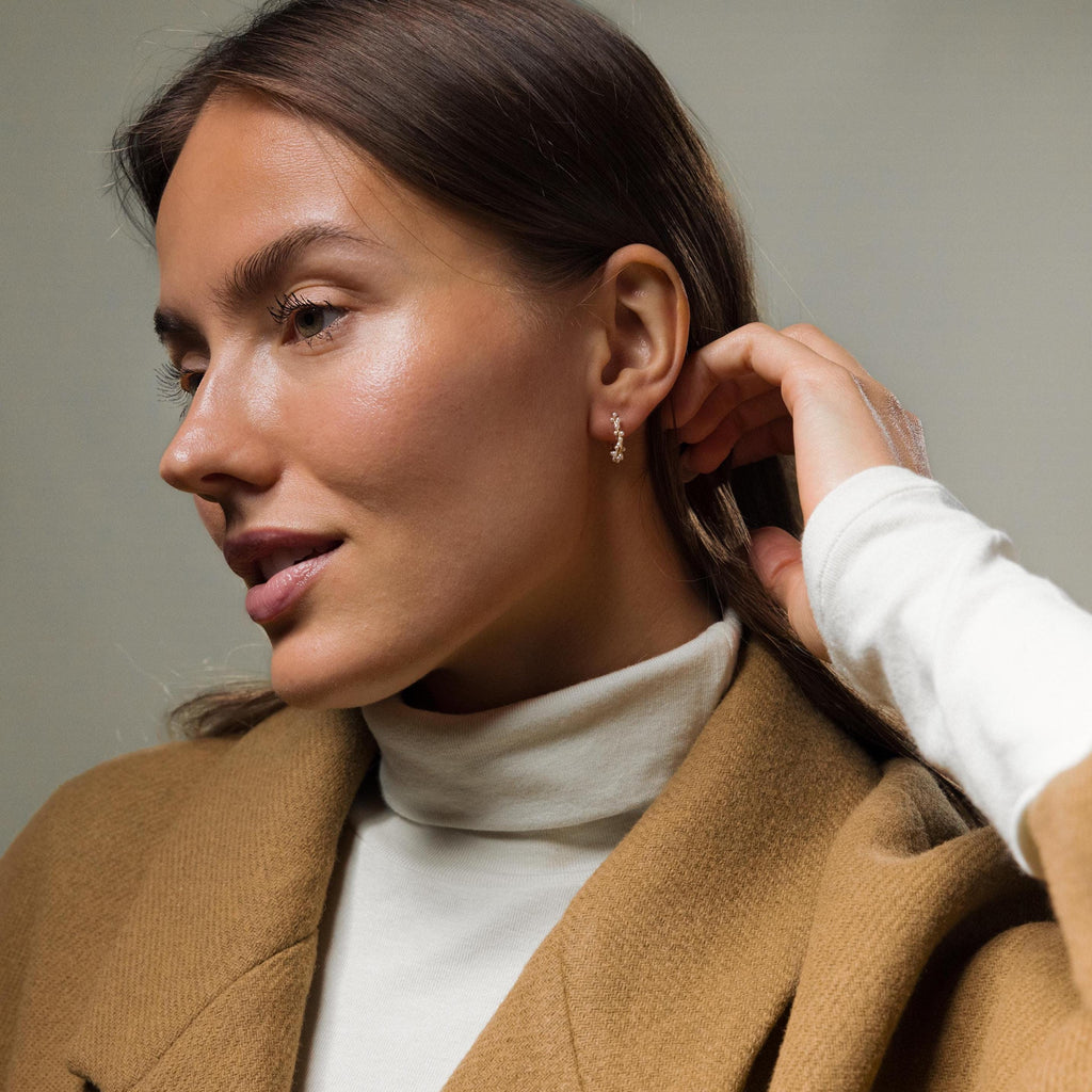 A woman in a tan coat and white turtleneck touches her Pearl Cluster Huggies in 18K Gold, gazing thoughtfully to the side.