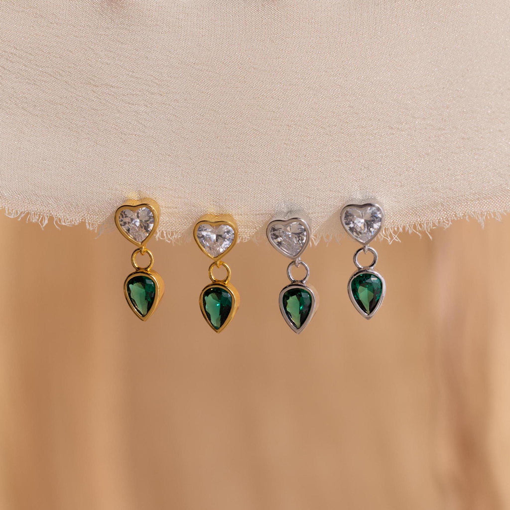 The Emerald Heart Drop Studs, along with three other heart and teardrop-shaped earrings in gold and silver finishes, are displayed elegantly on a light fabric.