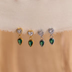 The Emerald Heart Drop Studs, along with three other heart and teardrop-shaped earrings in gold and silver finishes, are displayed elegantly on a light fabric.