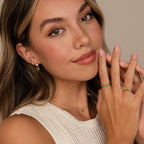 A woman with long hair smiles softly, wearing a sleeveless top and the Emerald Heart Drop Studs—elegant gold and green earrings that add a touch of glamour to her look.