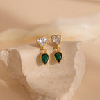 Emerald Heart Drop Studs feature heart-shaped clear stones and dangling emerald teardrop gems, elegantly displayed on a beige stone.