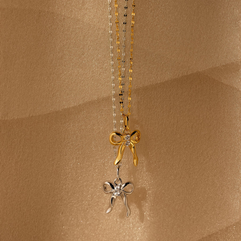 The Diamond Bow Charm in 18K Gold features a bow-shaped pendant adorned with sparkling CZ diamonds, adding coquette-inspired elegance to any look. Shown against a beige textured background for sophisticated appeal.