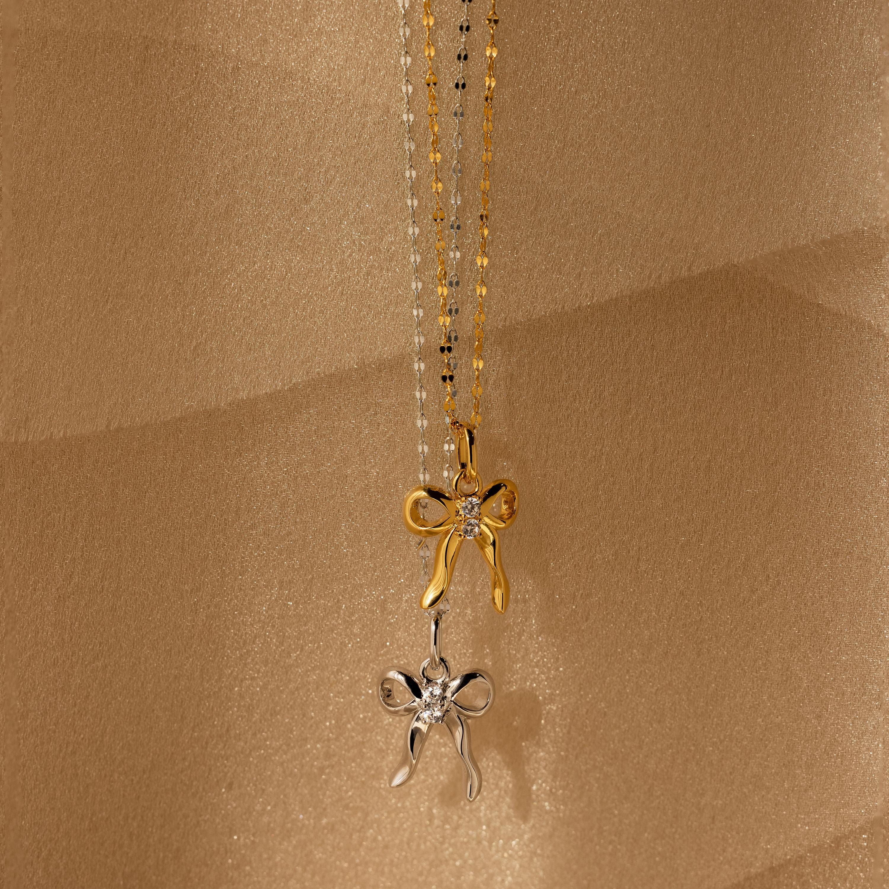 The Diamond Bow Charm in 18K Gold features a bow-shaped pendant adorned with sparkling CZ diamonds, adding coquette-inspired elegance to any look. Shown against a beige textured background for sophisticated appeal.
