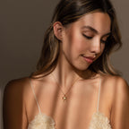 Woman with long hair wears a cream floral top and the Diamond Bow Charm in 18K Gold, looking down with a soft smile—a perfect coquette-inspired accessory moment.