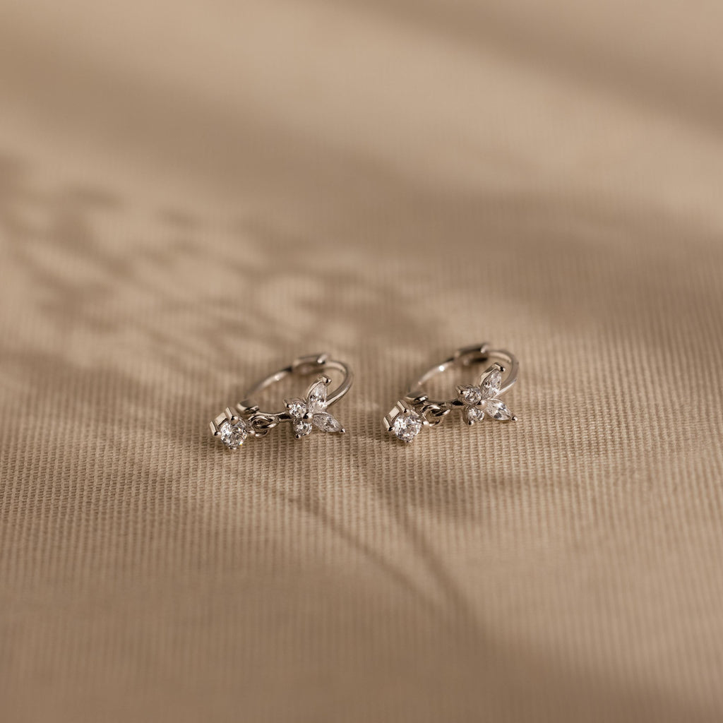 The Butterfly Drop Huggies in Sterling Silver, featuring delicate butterflies with small clear gemstones and diamond accents, rest on a beige textured surface.