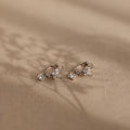 The Butterfly Drop Huggies in Sterling Silver, featuring delicate butterflies with small clear gemstones and diamond accents, rest on a beige textured surface.