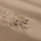 The Butterfly Drop Huggies in Sterling Silver, featuring delicate butterflies with small clear gemstones and diamond accents, rest on a beige textured surface.