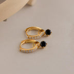 The Black Diamond Drop Huggies feature gold hoops with clear stones and a single black gemstone, displayed on a light beige surface. They’re perfect for adding subtle elegance and sophistication to any outfit.