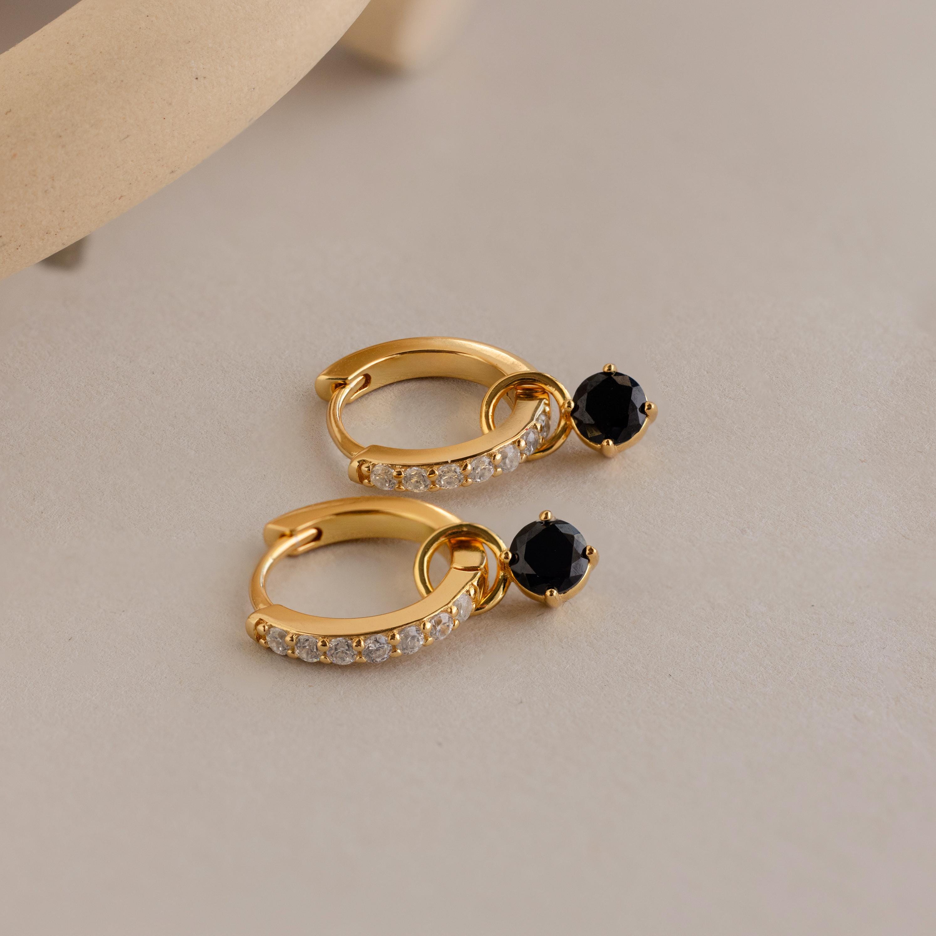 The Black Diamond Drop Huggies feature gold hoops with clear stones and a single black gemstone, displayed on a light beige surface. They’re perfect for adding subtle elegance and sophistication to any outfit.
