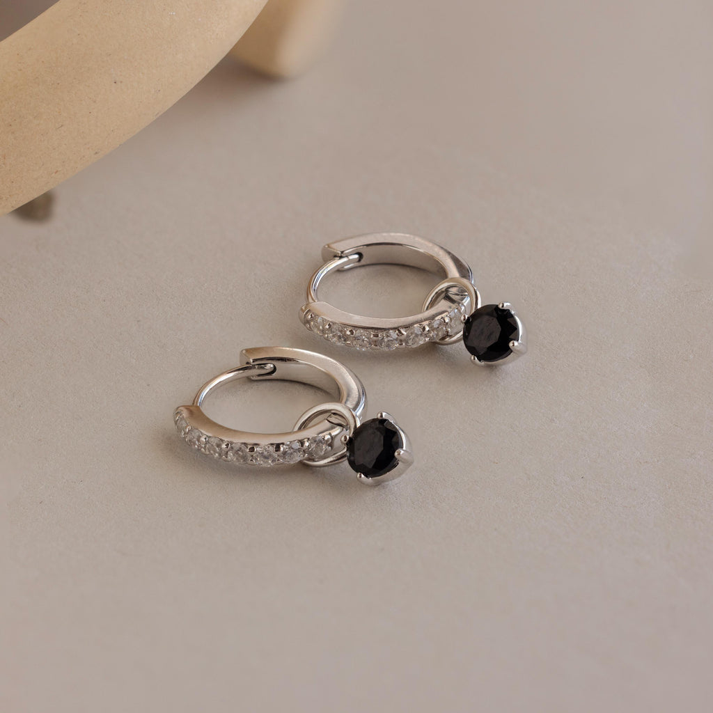 Silver hoop earrings with small black gemstone charms and clear stones, displayed on a neutral surface. The Black Diamond Drop Huggies offer a chic, modern touch with their elegant design and sparkling accents.