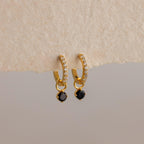 Black Diamond Drop Huggies: Elegant gold hoops featuring small clear stones and striking black gemstone dangles, showcased on a textured beige surface.
