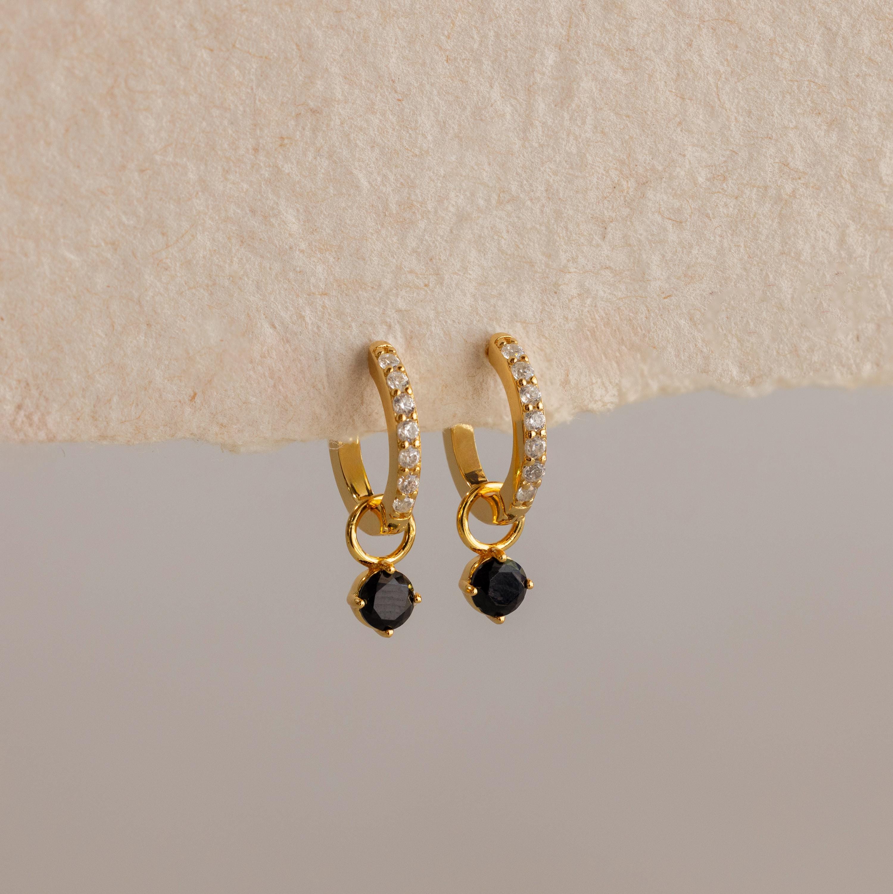 Black Diamond Drop Huggies: Elegant gold hoops featuring small clear stones and striking black gemstone dangles, showcased on a textured beige surface.