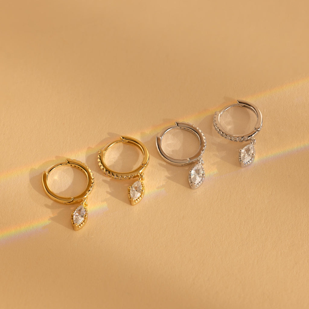 Two pairs of elegant earrings—one gold, one silver—feature a dangling marquise-cut gem, inspired by the Marquise Pave Drop Huggies, and are displayed on a beige surface.